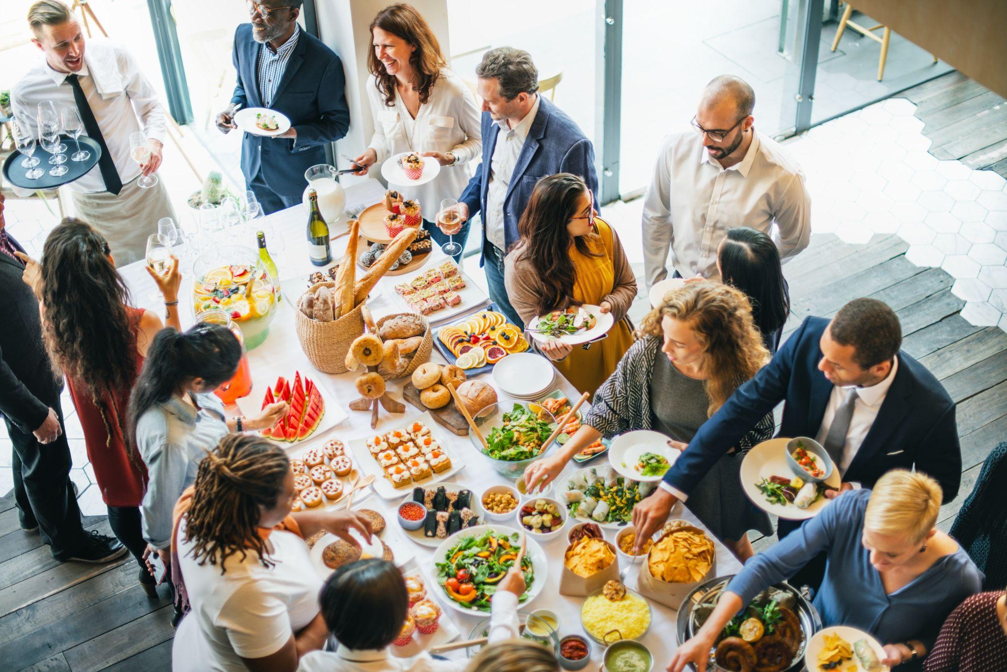 Overhead view of a diverse group of people serving themselves from a large buffet table filled with assorted foods and salads at a corporate event.